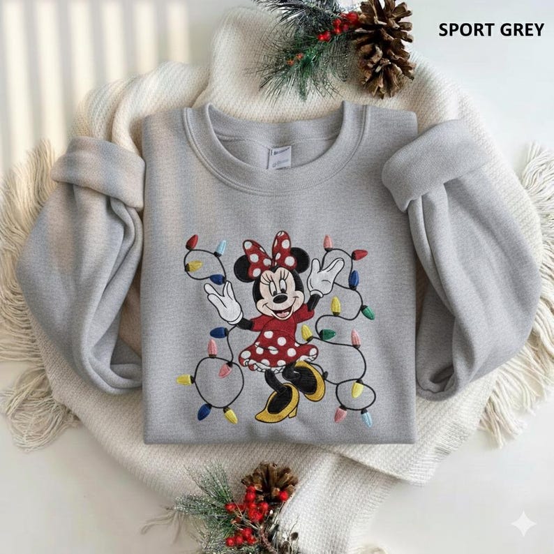 Op de afbeelding: Sportgrijs sweatshirt met een geborduurd Minnie Mouse-ontwerp. Minnie draagt een rode jurk met witte stippen en een bijpassende strik. Ze is omringd door kleurrijke kerstverlichting. De tekst "SPORT GREY" staat rechtsboven.