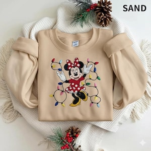 Op de afbeelding: Een zandkleurige sweatshirt met een geborduurd Minnie Mouse-ontwerp. Minnie draagt een rode jurk met witte stippen en een bijpassende strik. Ze is omringd door kleurrijke kerstverlichting. Het woord "SAND" staat rechtsboven.