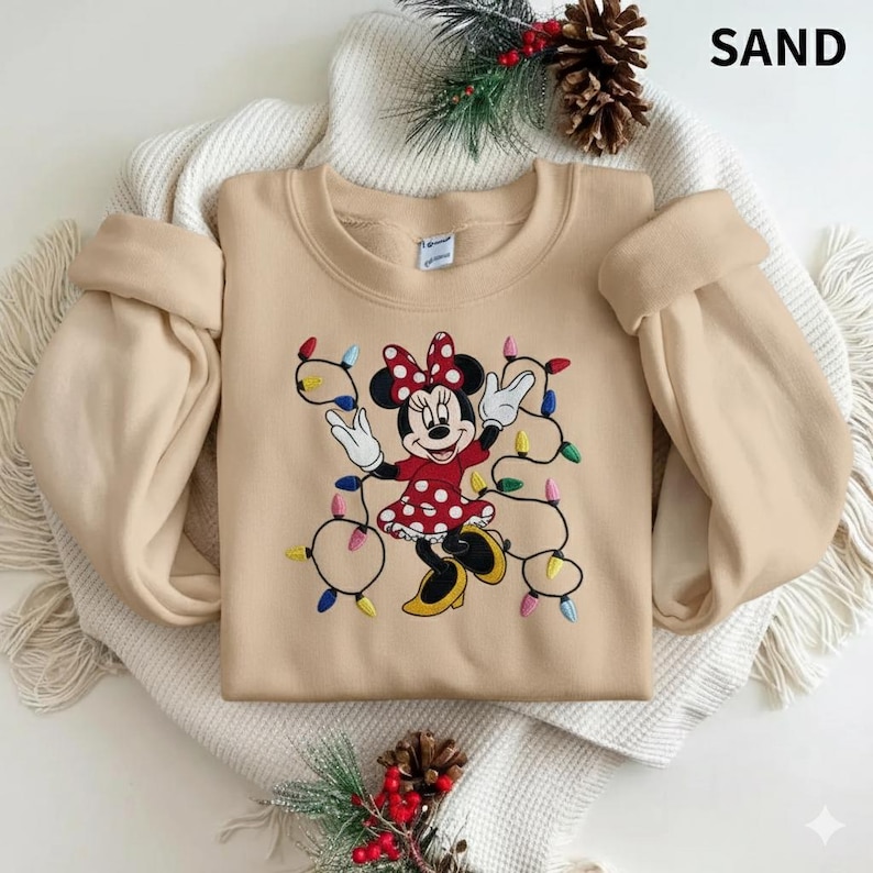 Op de afbeelding: Een zandkleurige sweatshirt met een geborduurd Minnie Mouse-ontwerp. Minnie draagt een rode jurk met witte stippen en een bijpassende strik. Ze is omringd door kleurrijke kerstverlichting. Het woord "SAND" staat rechtsboven.