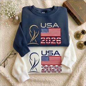 Puede incluir: Se muestran dos sudaderas, una azul marino y otra blanca. Cada una presenta el texto "USA" sobre una bandera estadounidense roja, blanca y azul y el número "2026". Un emblema dorado está en el lado izquierdo del diseño.