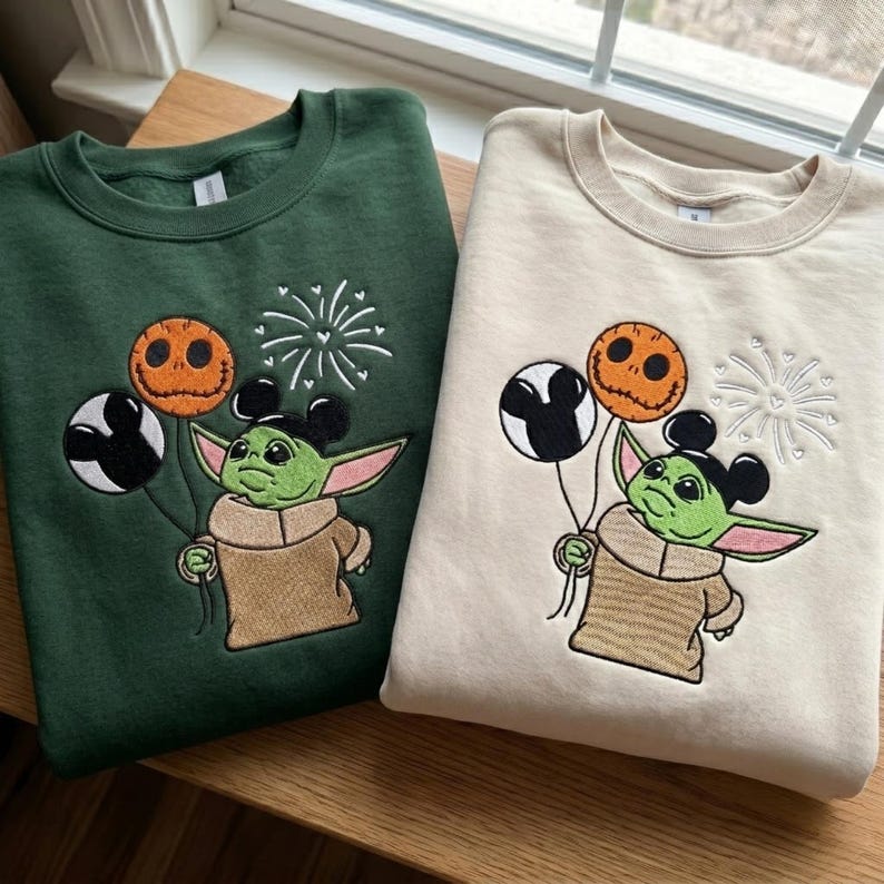 May include: A dark green sweatshirt and a cream sweatshirt, each featuring an embroidered graphic of a green creature with large ears wearing mouse-ear headbands, holding a black and white balloon and an orange pumpkin balloon with fireworks.