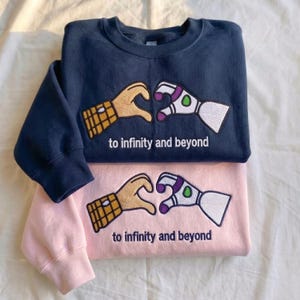 Puede incluir: Dos sudaderas, una azul marino y otra rosa, apiladas. Cada una presenta un dise&ntilde;o bordado de dos manos formando un coraz&oacute;n, una que se asemeja a un guante y la otra a un brazo rob&oacute;tico. El texto "to infinity and beyond" est&aacute; debajo.