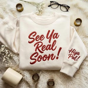 Może przedstawiać: Kremowa bluza z haftowanym napisem "See Ya Real Soon!" w czerwonym skrypcie z przodu. Prawy rękaw ma napis "Hiya Pal!". Bluza leży na puszystej białej powierzchni.