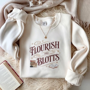 May include: Cream-colored sweatshirt with the words "Flourish and Blotts" in burgundy script, along with illustrations of books. A gold necklace with a pendant is visible. The sweatshirt is folded on a fluffy, cream-colored surface.