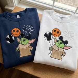 May include: A navy blue sweatshirt and a white sweatshirt, each featuring an embroidered green creature with large ears wearing a tan robe. The creature holds three balloons: one black and white Mickey-shaped, one orange Jack Skellington-style, and a firework design.