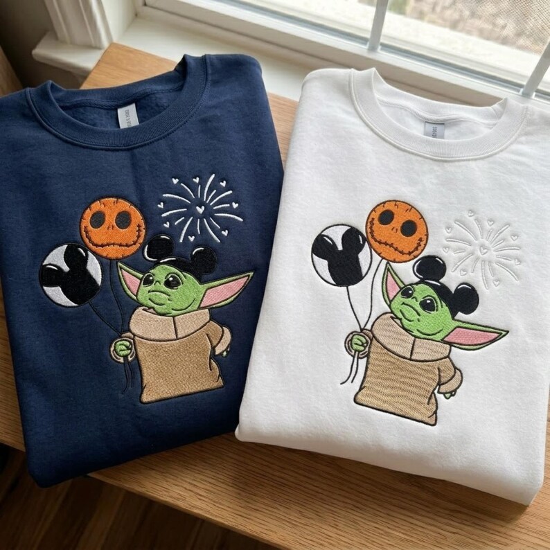 May include: A navy blue sweatshirt and a white sweatshirt, each featuring an embroidered green creature with large ears wearing a tan robe. The creature holds three balloons: one black and white Mickey-shaped, one orange Jack Skellington-style, and a firework design.