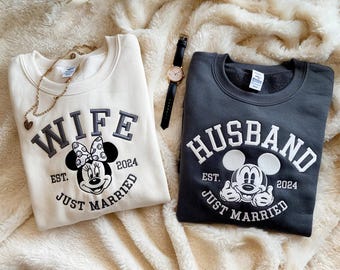 May include: Two matching sweatshirts, one cream and one charcoal, with "WIFE" and "HUSBAND" in white block letters. Each features a Mickey Mouse design and the text "EST. 2024 JUST MARRIED". A gold necklace and a watch with a black band are also visible.