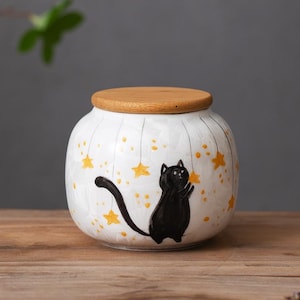 Pode incluir: Jarro de cerâmica branca com tampa de madeira, apresentando um gato preto alcançando uma estrela amarela. O jarro é decorado com um padrão de estrelas amarelas sobre um fundo branco.