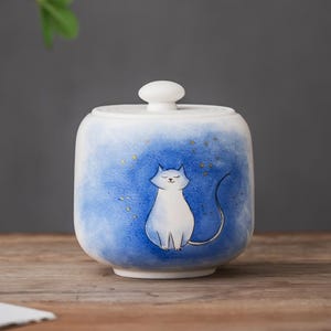 May include: A white ceramic jar with a lid, decorated with a hand-painted white cat on a blue watercolour background. The jar has a rounded shape and a small, white knob. Gold flecks add a touch of sparkle. The jar is on a wooden surface.