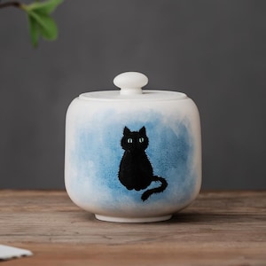 Puede incluir: Un tarro de cerámica blanco con tapa, con un gato negro pintado a mano sobre un fondo azul acuarela. El tarro tiene forma cuadrada redondeada y un pequeño pomo redondo en la parte superior.