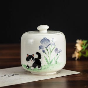 May include: A white ceramic jar with a lid, decorated with a hand-painted design of a black and white kitten and blue iris flowers. The jar is on a piece of paper, with a dark background. A decorative item.
