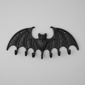 May include: A black metal bat-shaped wall hook with six hooks. The bat has detailed wings and a smiling face. The wings are textured with a ribbed pattern. A decorative and functional item, perfect for Halloween or gothic-themed interiors.