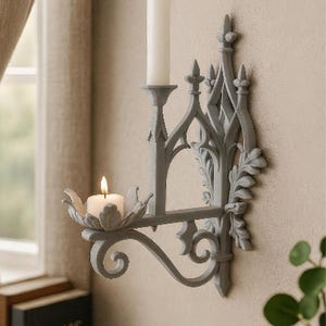 Puede incluir: Un candelabro de pared gris con un diseño arquitectónico intrincado de arcos apuntados, motivos de hojas y dos plataformas. La plataforma superior sostiene una vela blanca alta, mientras que la inferior sostiene una pequeña vela de té.