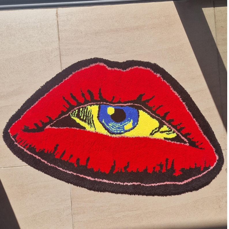 May include: A red rug shaped like a mouth with an eye design. The lips are bright red, outlined in black, and the eye features a blue iris, yellow iris, and black details. The rug is a unique home decor item.