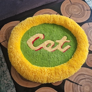 May include: A round, handmade cat bed with a yellow border and a green center. The word "Cat" is embroidered in the center in a cream color with red outlines. The bed is made of a soft, textured material.