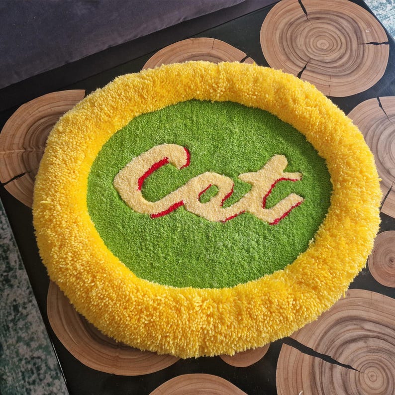 May include: A round, handmade cat bed with a yellow border and a green center. The word "Cat" is embroidered in the center in a cream color with red outlines. The bed is made of a soft, textured material.