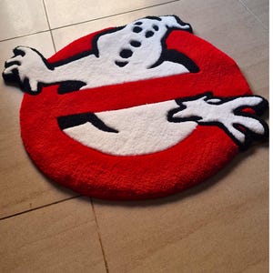 May include: A round, red rug featuring the Ghostbusters logo. The logo includes a white ghost figure with black accents, set against a red circle with a white slash through it. The rug is made of a soft, plush material.
