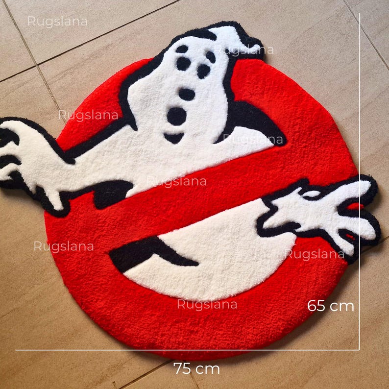 May include: A round, red rug featuring the Ghostbusters logo. The rug has a white ghost figure with black details inside a red circle with a white slash. The rug measures 75 cm by 65 cm.
