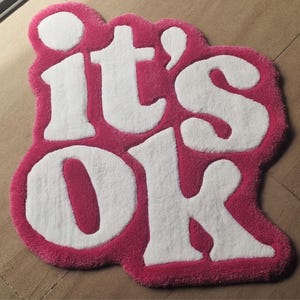 May include: A pink and white rug with the words "it's OK" in a playful, bubble-like font. The rug has a soft, plush texture and a unique, cloud-like shape. The letters are white against a pink background.