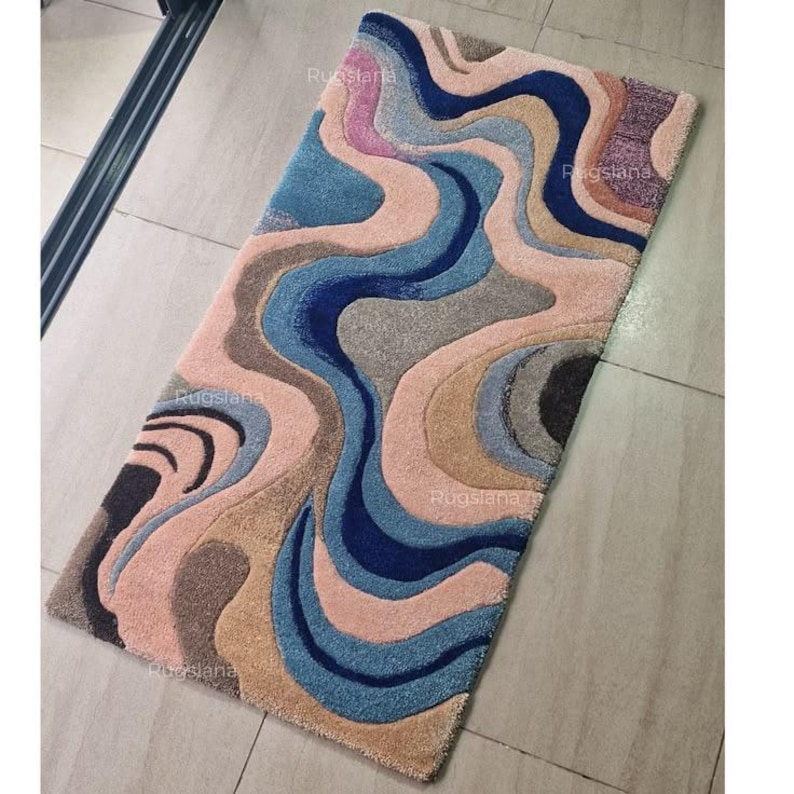 May include: A rectangular rug with a flowing, abstract design. The rug features a mix of colors, including shades of blue, pink, gray, and brown. The pattern resembles water currents or layered textures. The rug is made of a soft, plush material.