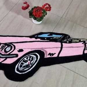 May include: A pink car-shaped rug with black trim and wheels. The rug features a classic car design with a convertible top. A small white pot with red flowers sits in the background.