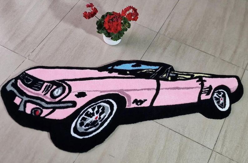 May include: A pink car-shaped rug with black trim and wheels. The rug features a classic car design with a convertible top. A small white pot with red flowers sits in the background.