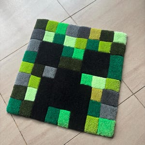 May include: A square, handmade rug featuring a pixelated design of a Creeper from the video game Minecraft. The rug is primarily black, green, and gray, with a soft, textured surface. The rug is designed to be placed on the floor.