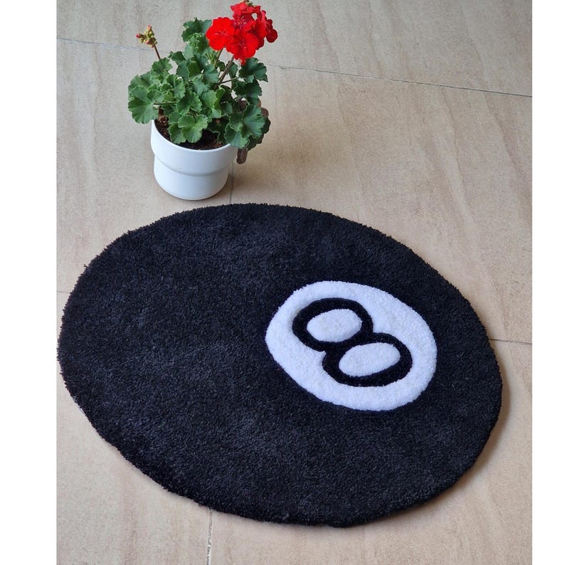 May include: Black, oval-shaped bath mat with a white number eight design. The mat is made of a soft, plush material. A potted plant with red flowers sits in the background. The mat is designed to resemble a pool ball.