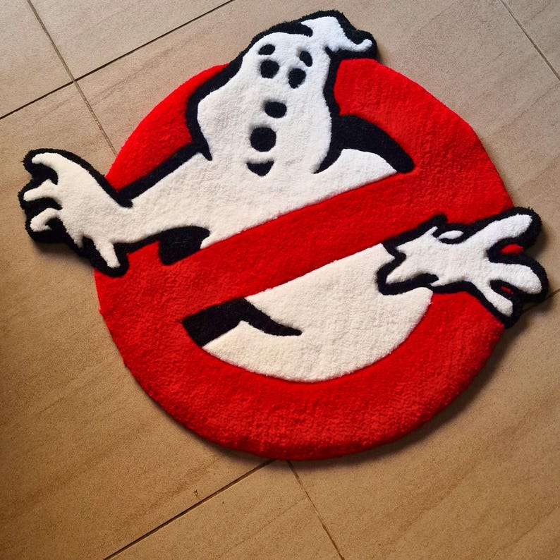 May include: A round, red, white, and black rug featuring the Ghostbusters logo. The rug has a white ghost figure with black eyes and mouth, and white hands. The ghost figure is set against a red circle with a white slash through it.