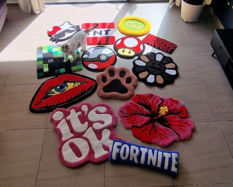 May include: A collection of colorful, decorative rugs in various shapes and designs. The rugs include a red lip, a paw print, a flower, a mushroom, and the text "Fortnite". A white cat is standing on the rugs.