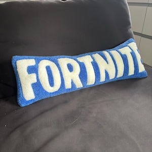 May include: A rectangular blue pillow with the word "FORTNITE" in large white letters. The pillow is made of a soft, textured material and is placed on a dark gray surface.