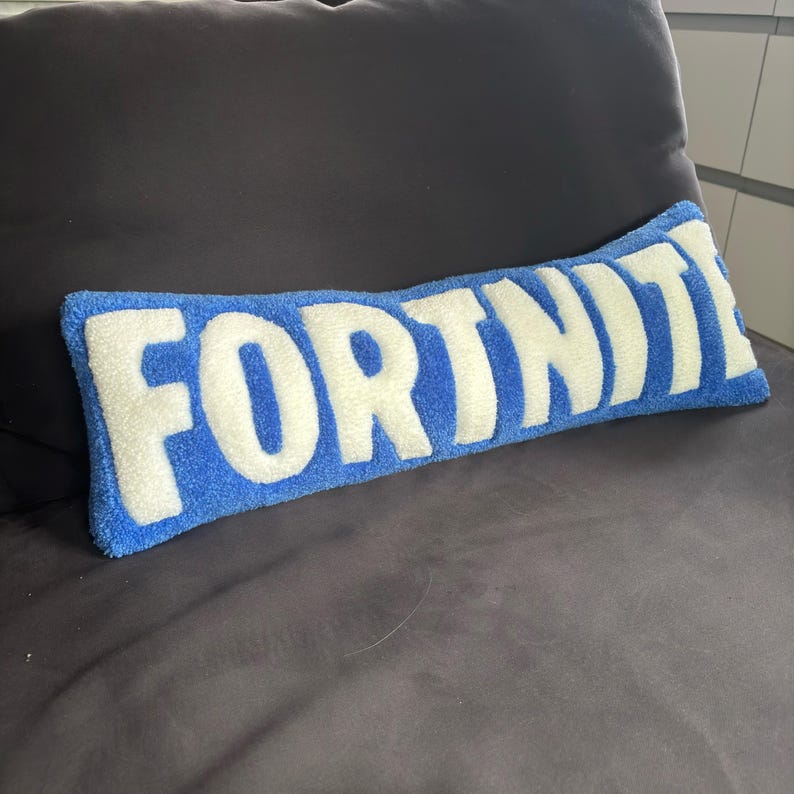 May include: A rectangular blue pillow with the word "FORTNITE" in large white letters. The pillow is made of a soft, textured material and is placed on a dark gray surface.
