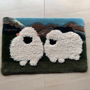 May include: A rectangular rug with two sheep with fluffy, cream-coloured wool and black eye details. The background features a green field, brown hills, and a blue sky. The rug has a textured finish, with the brand name "RugsLana" visible.
