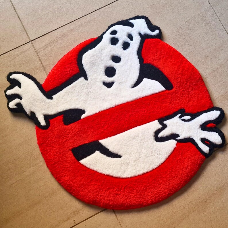 May include: A round, red and white rug featuring the Ghostbusters logo. The rug has a red circle with a white ghost figure and a black no-ghost symbol. The rug is made of a soft, plush material.