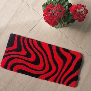 May include: A rectangular rug with a wavy pattern of red and black stripes. The rug is made of a soft, textured material. A potted plant with red flowers is in the background.
