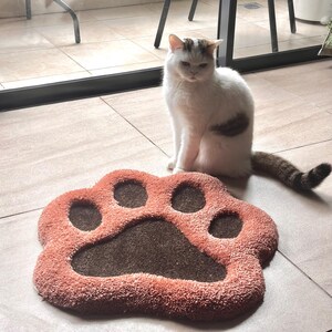 May include: A plush, paw-shaped pet mat in shades of coral and brown. A white and brown cat sits nearby. The mat has a textured surface and is designed to resemble a large animal paw print.