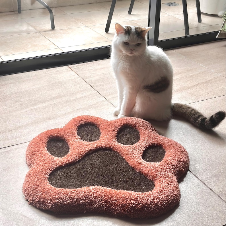 May include: A plush, paw-shaped pet mat in shades of coral and brown. A white and brown cat sits nearby. The mat has a textured surface and is designed to resemble a large animal paw print.