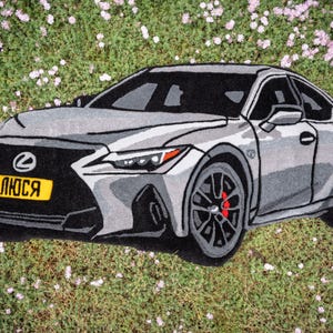 May include: A gray and black car rug with a license plate that reads "ЛЮСЯ" in yellow. The car rug is on a bed of green grass with small pink flowers.
