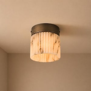 Op de afbeelding: Een plafondlamp met een cilindrische kap van geribbelde, doorschijnende witte steen met grijze aders. Het armatuur is bevestigd met een ronde basis van donkerbronskleurig metaal die aan het plafondoppervlak is gemonteerd.