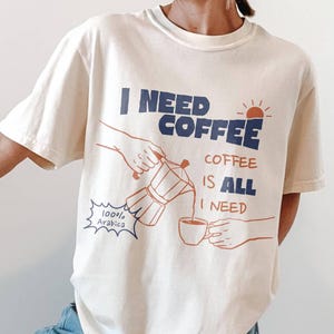 May include: Cream-colored t-shirt with the text "I NEED COFFEE COFFEE IS ALL I NEED" in navy blue and red. The graphic depicts a coffee pot pouring coffee into a cup. The person is wearing light blue jeans and white sneakers.