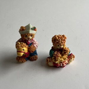 May include: Two small teddy bear figurines. One is wearing a sailor hat and holding a yellow rocking horse. The other is wearing a blue and red dress and holding a red and white gift.
