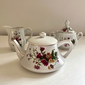 May include: A white porcelain tea set with a floral design. The set includes a teapot, creamer, and sugar bowl.