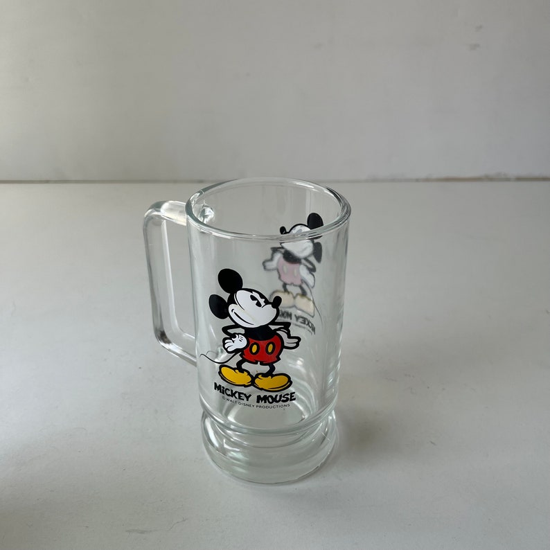 May include: A clear glass mug with a handle features a black and white cartoon image of Mickey Mouse wearing red shorts and yellow shoes. The mug has the text "Mickey Mouse" and "Walt Disney Productions" printed on the bottom.