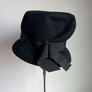 May include: A black wool hat with a wide brim and a ribbon band. The hat features a large bow on the side. The hat is displayed on a metal stand against a white background. The hat's design is classic and elegant.