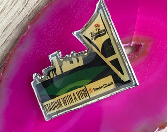 May include: A silver pin with a green, white, and black image of a stadium with the text "Stadium with a View" and "RadioShack" on it. The pin also has a red and white image of a baseball team logo with the text "Yankees" on it.