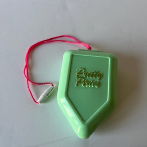 May include: A light green, shield-shaped compact with the words "Pretty Pixies" in gold script. It has a pink cord attached with a white plastic piece. The compact is closed and appears to be made of plastic.