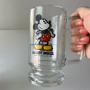 May include: A clear glass mug with a Mickey Mouse illustration. Mickey is wearing his signature red shorts and yellow shoes. The mug has a handle and a rounded base. The text "Mickey Mouse" and "&copy; WALT DISNEY PRODUCTIONS" is printed on the mug.