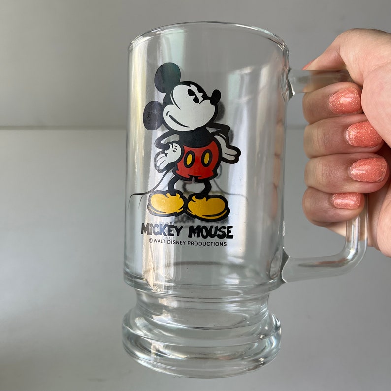 May include: A clear glass mug with a Mickey Mouse illustration. Mickey is wearing his signature red shorts and yellow shoes. The mug has a handle and a rounded base. The text "Mickey Mouse" and "&copy; WALT DISNEY PRODUCTIONS" is printed on the mug.