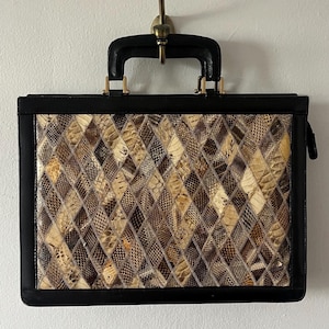 May include: A rectangular briefcase featuring a central panel with a diamond-patterned mosaic of textured snakeskin in shades of brown, tan, and cream. The frame, edges, and top handle are constructed from smooth black material with gold-tone hardware.