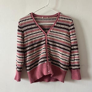 May include: A V-neck cardigan sweater with a button-up front. The sweater features a pattern of horizontal stripes in shades of pink, white, brown, and black. The cuffs and bottom hem are solid pink. The sweater is hanging on a white hanger.